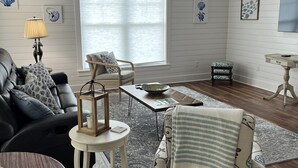 Un salón amplio con techo alto, un ventanal grande y chimenea. Hay un sofá de cuero negro, un sillón estampado y una mesa redonda de madera.