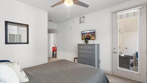 Ein Schlafzimmer mit Bett, Kommode, Deckenventilator, Fernseher und einer Tür, die auf einen Balkon führt.