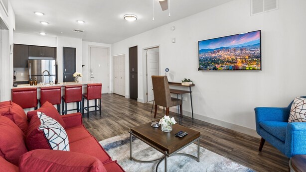 Ein modernes Wohnzimmer mit einem roten Sofa, einem blauen Sessel und einem Essbereich mit roten Barhocker. An der Wand hängt ein Stadtbild.
