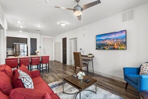 A modern living room with a red sofa, a blue armchair, and a dining area with red bar stools. A cityscape painting adorns the wall.