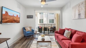 Ein Wohnzimmer mit einem roten Sofa, einem blauen Sessel und einem Couchtisch mit einer Vase. Es gibt einen Deckenventilator, ein großes Fenster und ein Gemälde an der Wand.