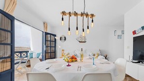 A dining area with a white table set for four, a chandelier, and a view of the sea through a large window.