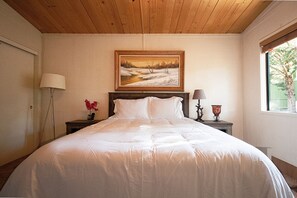 Un dormitorio con una cama grande, un cuadro sobre la cama, una mesita de noche con una lámpara y una ventana con vistas a árboles.