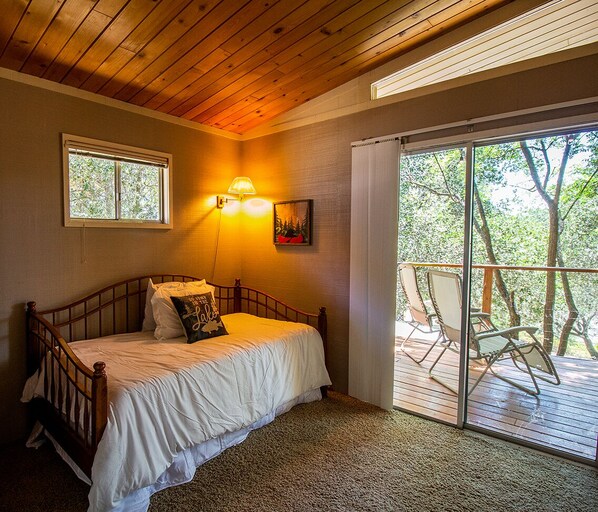 Un dormitorio con cama de madera, mesita de noche, lámpara y ventana. Tiene un balcón con sillas y vistas a árboles.
