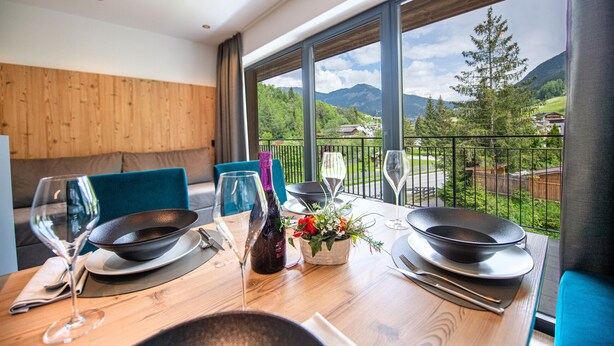 Comedor con mesa de madera dispuesta para dos personas, vista a las montañas y un balcón con barandilla.