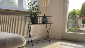 A room with a black coffee table displaying brochures, a beige sofa, a radiator, a window with decorative iron bars, and a potted plant. - Ferienwohnung Heidi (Rielasingen-Worblingen)