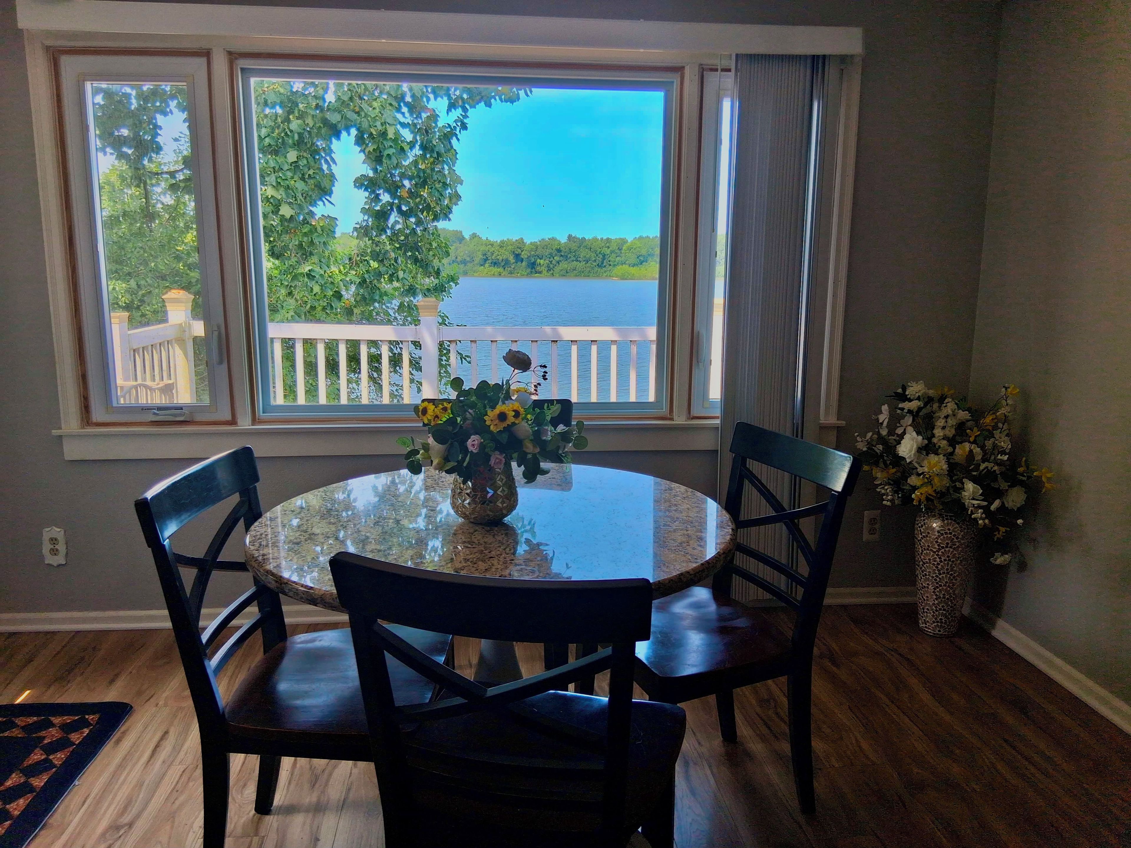 Un coin repas avec une table ronde, quatre chaises, un vase de fleurs et une vue sur un lac, visible à travers de grandes fenêtres.
