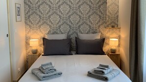 Un lit bien fait, avec un papier peint à motifs gris et blancs, deux lampes de chevet et une petite étagère sur laquelle est posée une lampe.