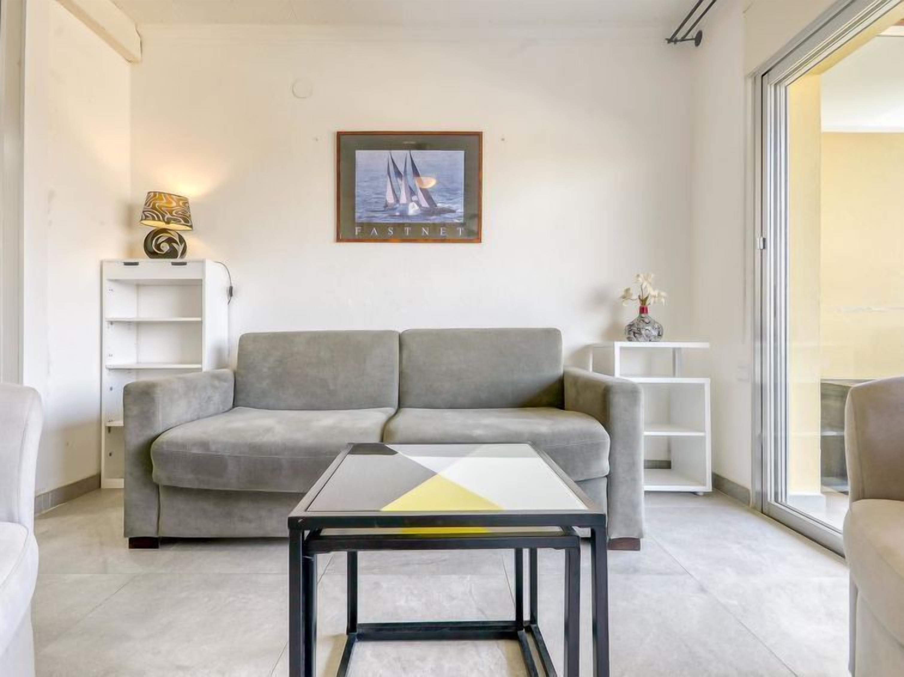 Een moderne woonkamer met een grijze bank, een salontafel met glazen blad en een ingelijst kunstwerk aan de muur.