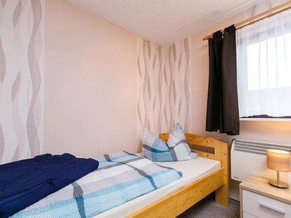 A single bed with a plaid blanket, a wooden headboard, a bedside table with a lamp, and a radiator. - Vacation home Ehmke by Interhome (Karlshagen)