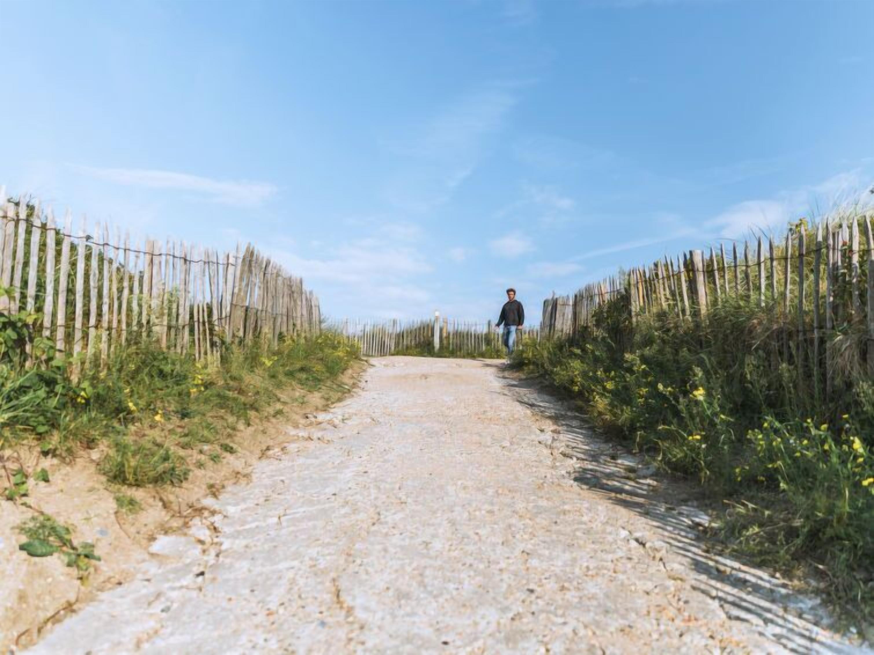 A dirt path flanked by wooden fences leading to a person walking away.