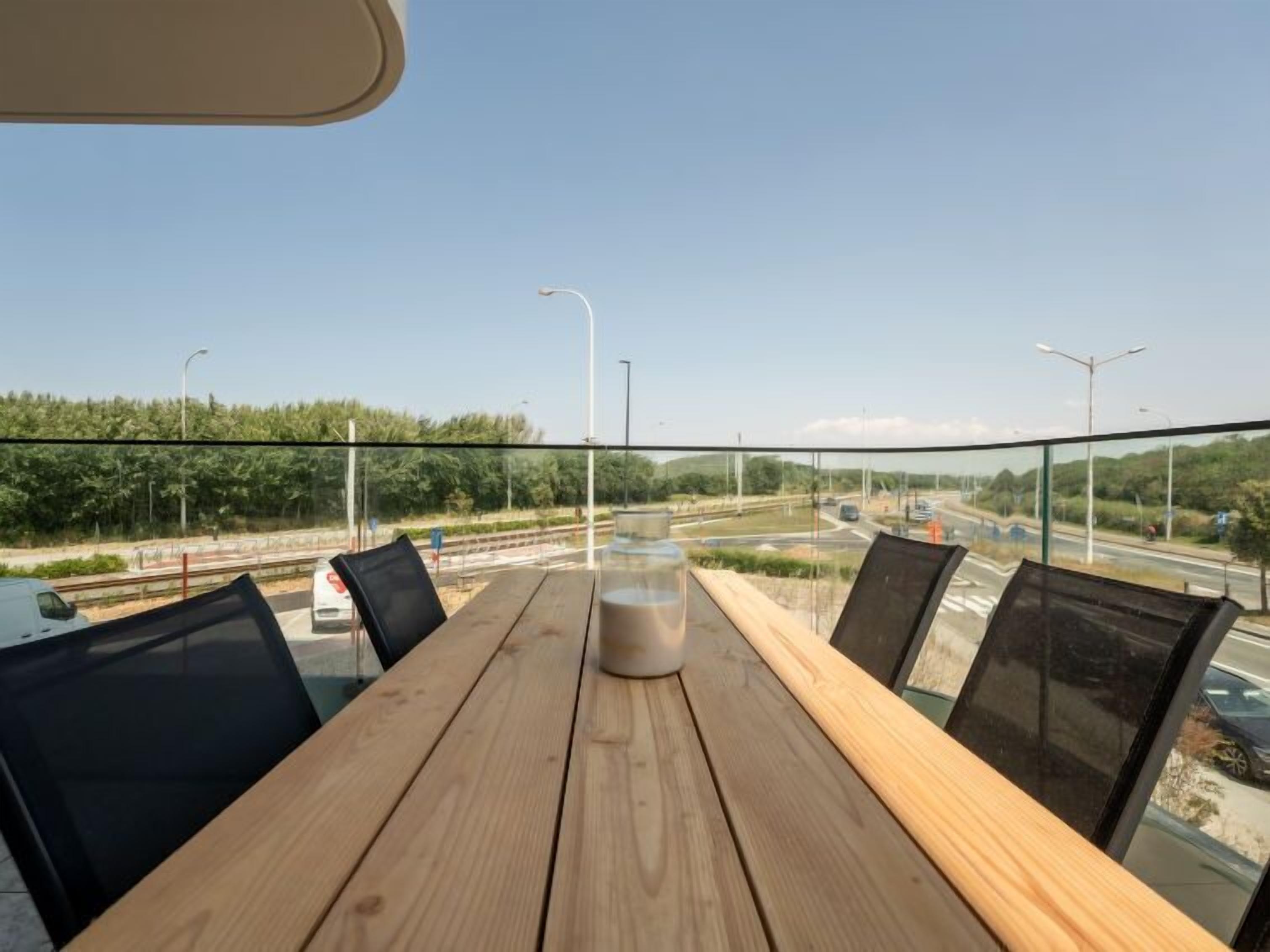 Una mesa de madera con sillas en una habitación con pared de vidrio que da a una autopista y follaje.