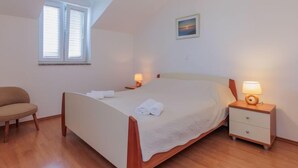 Un dormitorio con cama, mesitas de noche, una silla, una ventana y un cuadro en la pared.