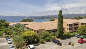 A coastal residential area with buildings featuring terracotta roofs, a parking lot, and a view of the sea and distant mountains.