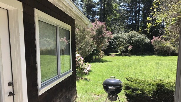 Une terrasse en bois mène à une maison dont le porche est agrémenté d’un barbecue noir.