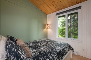Ein Schlafzimmer mit einem Bett, einem Nachttisch, einer Lampe und einem Fenster mit Blick auf Bäume.