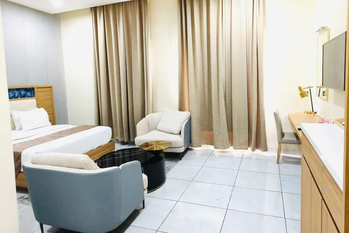 Una habitación de hotel con una cama, dos sillones, una mesita, un escritorio con espejo y cortinas.