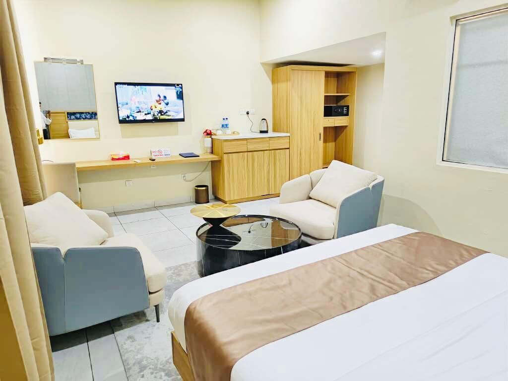 A hotel room with a bed, two armchairs, a small round table, a TV, a wooden cabinet, and a window.