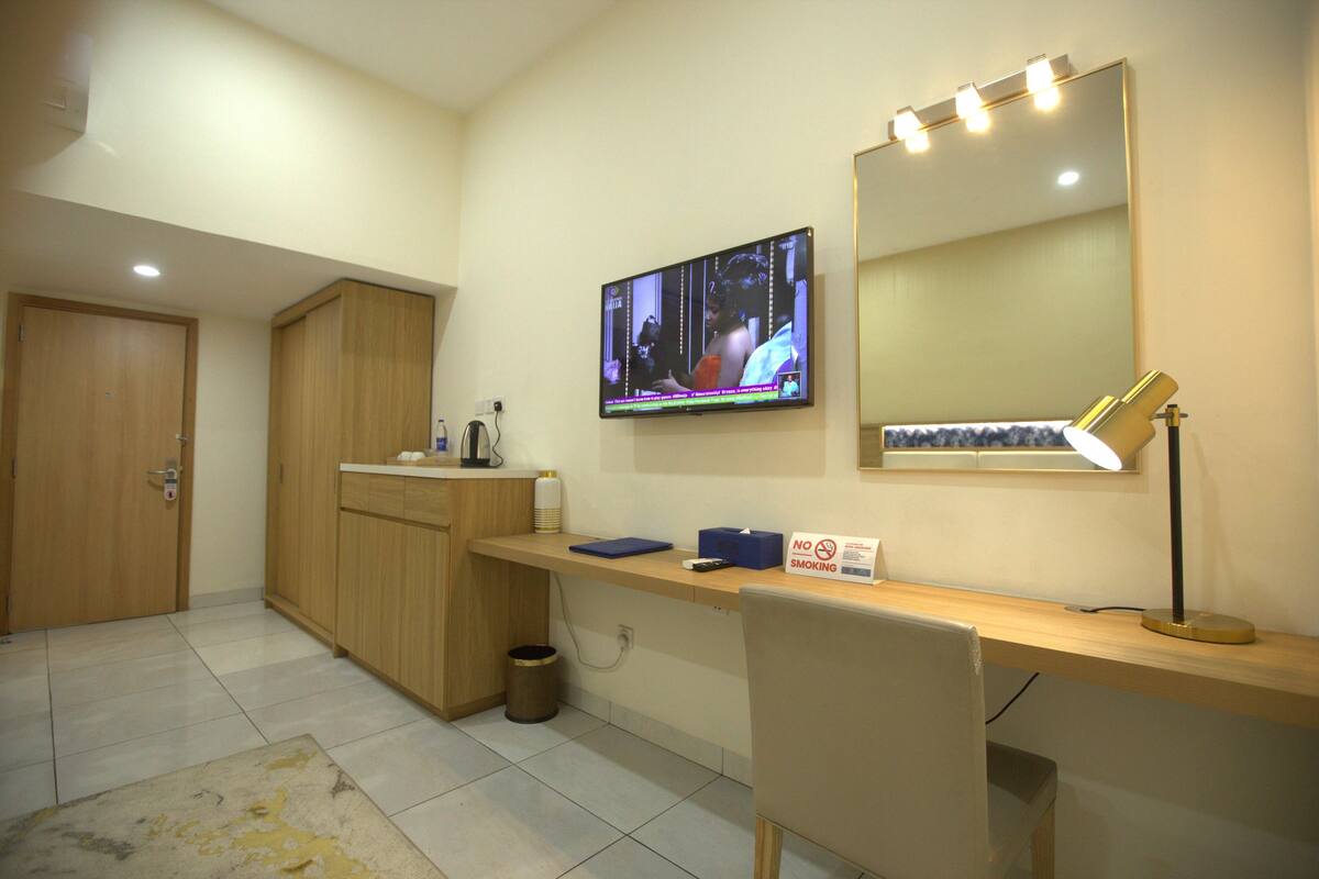 Habitación de hotel con un armario de madera, un escritorio con una computadora, un televisor colocado sobre el escritorio y un espejo con iluminación.