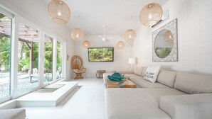 Una sala de estar moderna con un sofá blanco, televisión, lámparas colgantes de mimbre y un ventanal con vistas a un jardín.