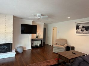 A living room with a fireplace, a television mounted on the wall, a ceiling fan, and a framed picture on the wall. - Cozy 3bdrm 2bath house for families w/ small kids & infants near White Rock Lake (Dallas)
