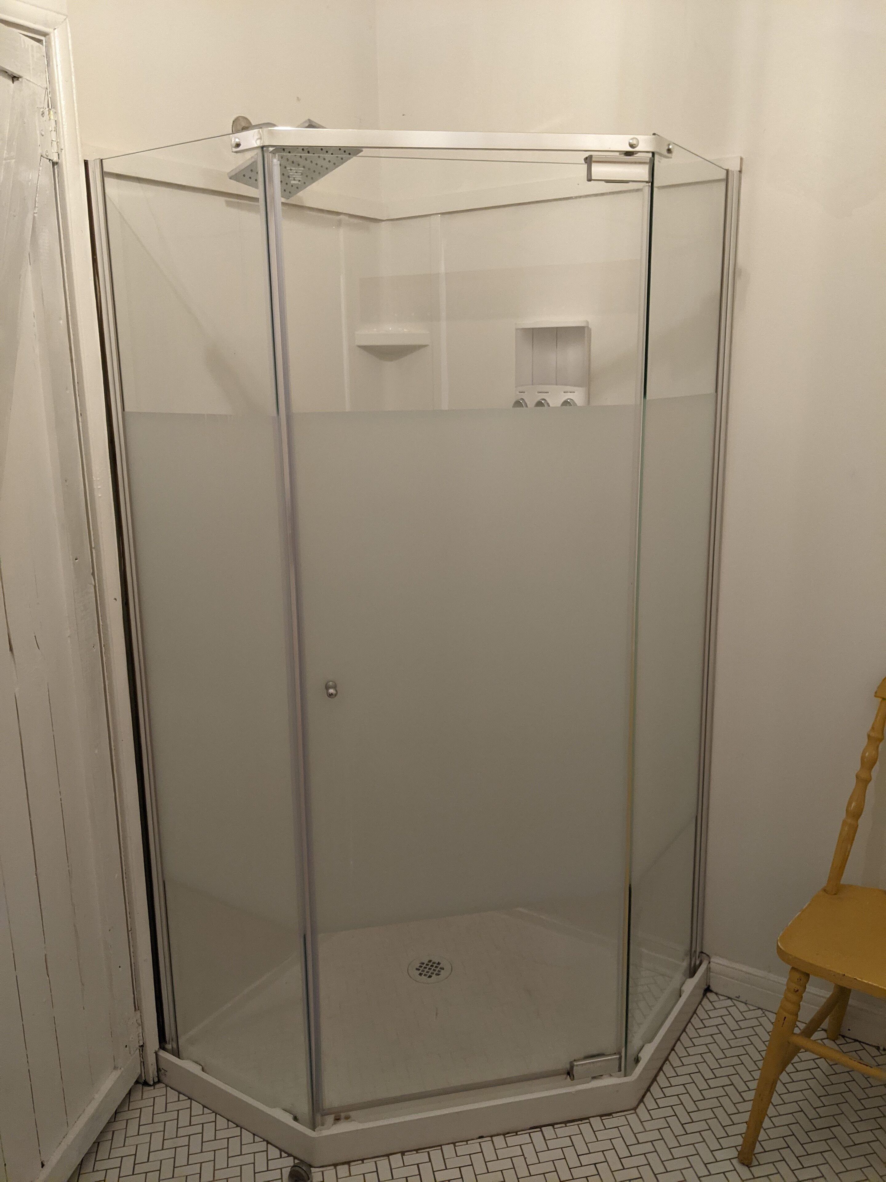 A glass-enclosed shower stall with a white tiled floor and a wooden chair in the corner.