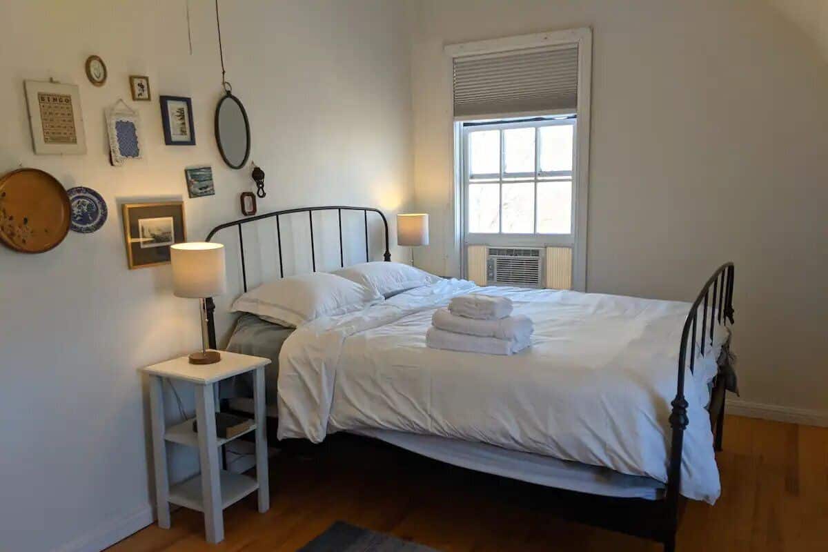 A neatly made bed with white linens, a bedside table with a lamp, and a window with blinds.