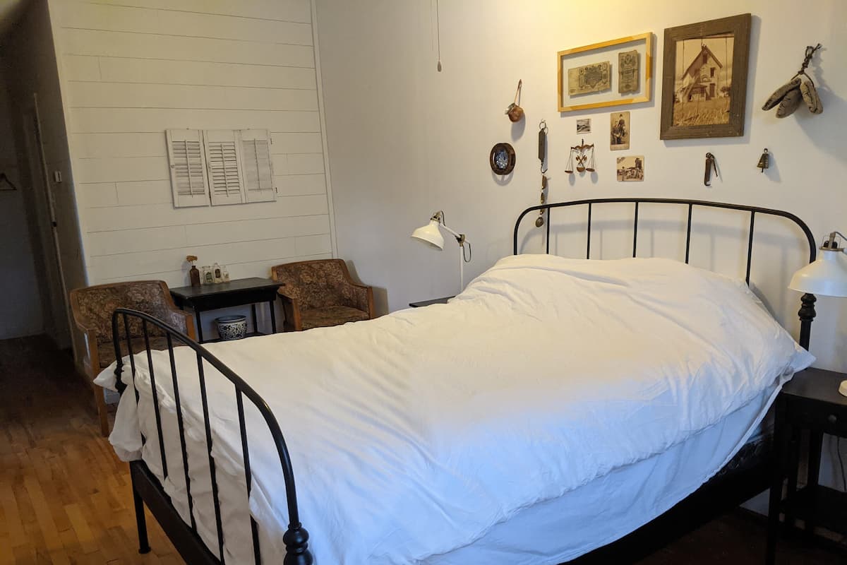 A single bed with a metal frame, a wooden chair, a small table, and a wall with framed pictures and various items.