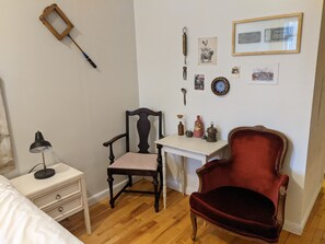 Ein Zimmer mit Holzboden, einem Schreibtisch, einem Stuhl, einem Bett und einer Wand mit verschiedenen Gegenständen, darunter eine Lampe, ein Löffel und gerahmte Bilder.