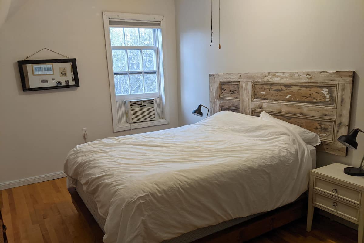 A bedroom with a wooden headboard, a white bedspread, a bedside table with a lamp, and a window with blinds.
