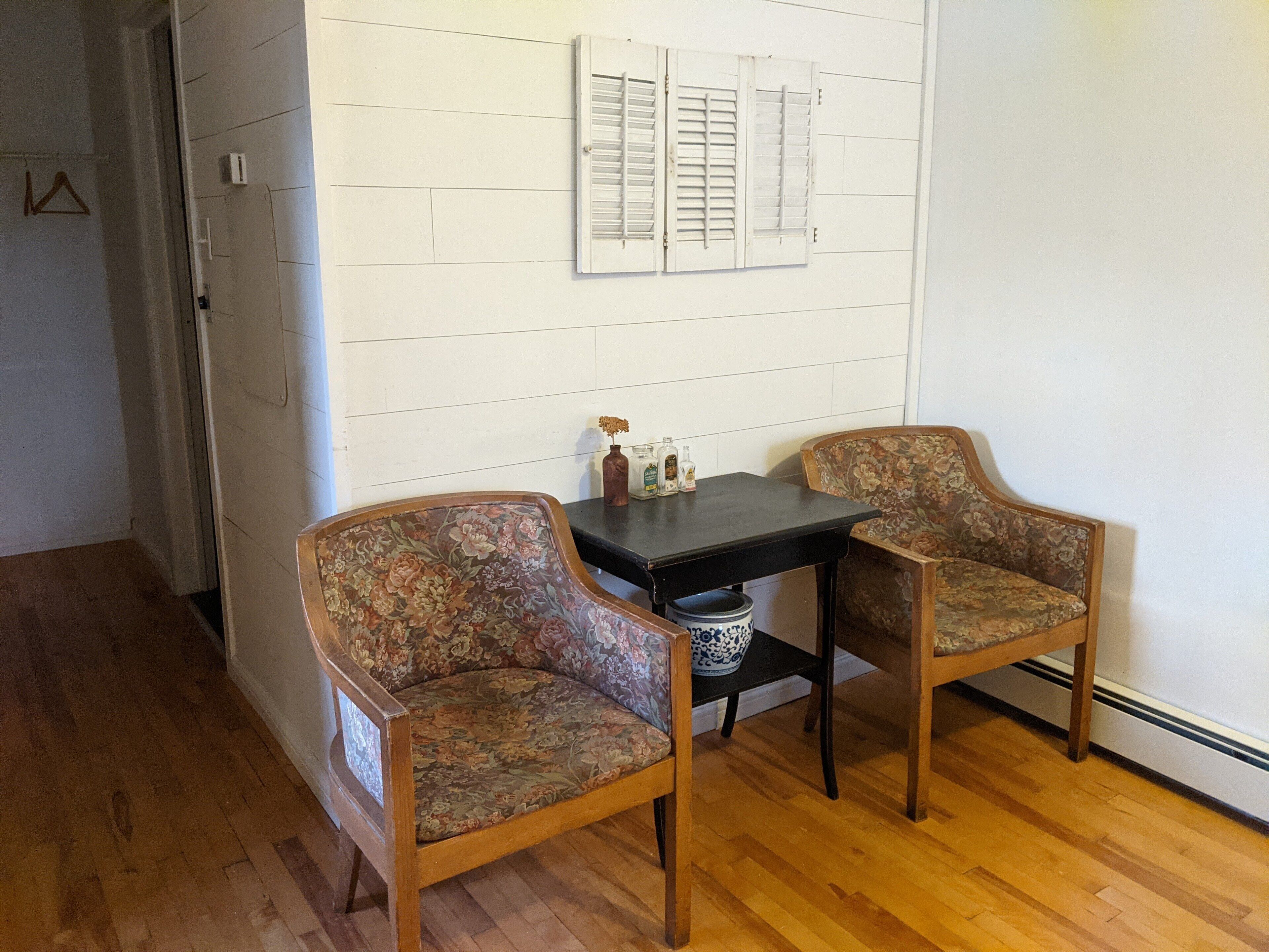 Una habitación con dos sillones de estampado floral, una mesita y un estante en la pared con objetos decorativos.