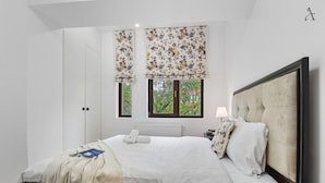 A bedroom with a large bed, a nightstand, a lamp, and two windows with floral blinds. - 1BDR Apartment close to Unirii Square (Bucharest)