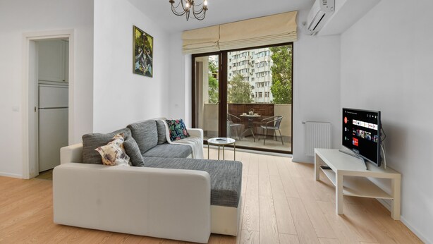 A modern living room with a sofa, a TV, and a balcony with a view of buildings and trees. - 1BDR Apartment close to Unirii Square (Bucharest)