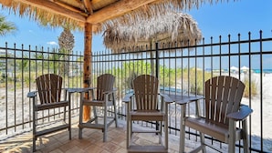 A covered outdoor seating area with wooden chairs and tables, a thatched roof, and a view of the beach.
