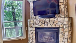 A stone fireplace with a television above it, a window with a view of trees, and a power outlet on the wall. - Chateau Treehouse #11/ Oak Crest (Eureka Springs)