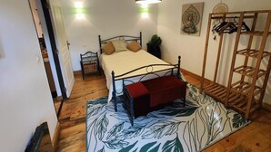 Une chambre à coucher comprenant un lit, des tables de chevet, un luminaire suspendu, un plancher en bois, une étagère fixée au mur et une porte donnant sur une autre pièce.