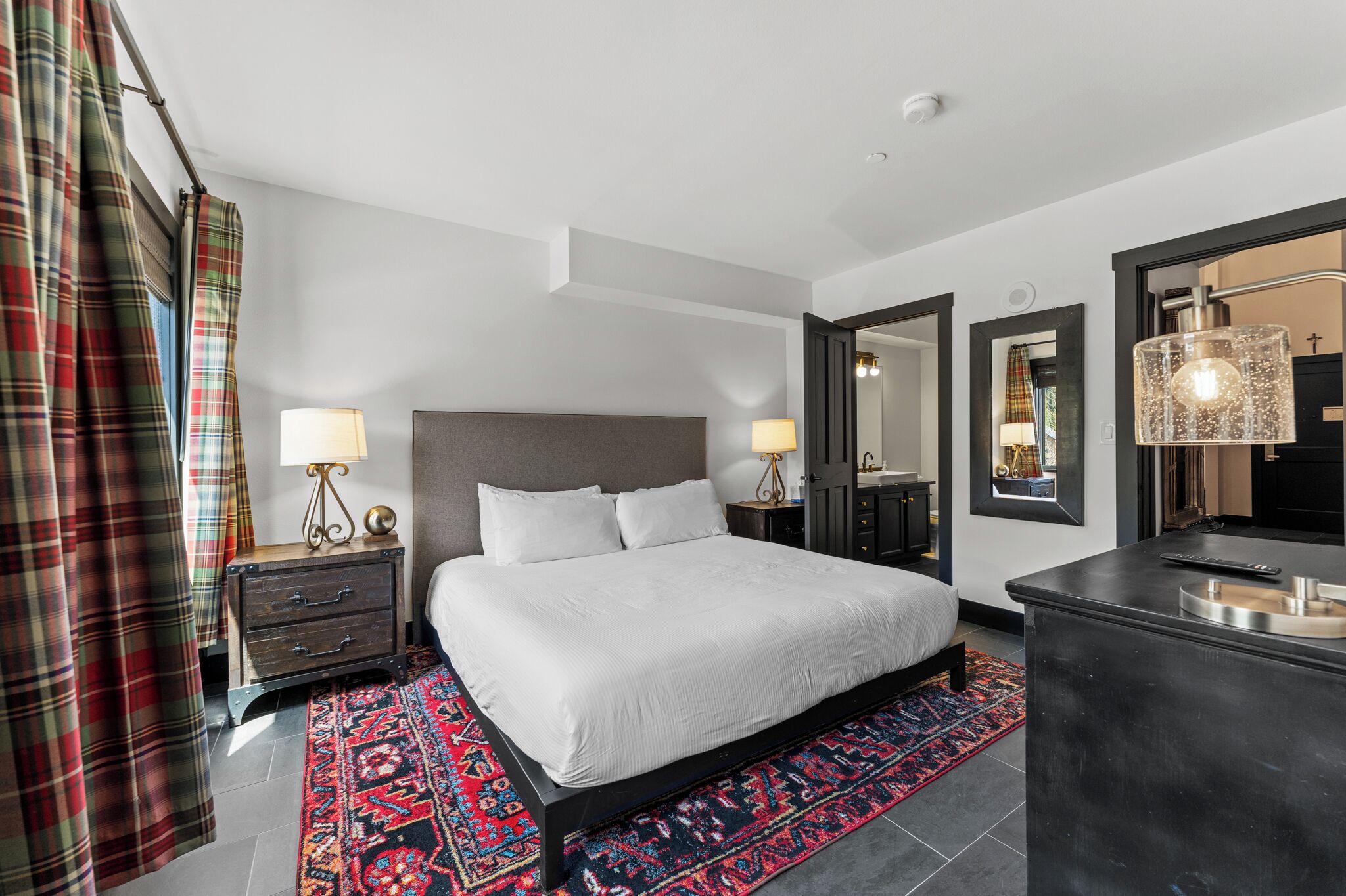 A modern hotel room with a large bed, two bedside tables with lamps, a dark headboard, a large mirror, and a small desk with a mirror.