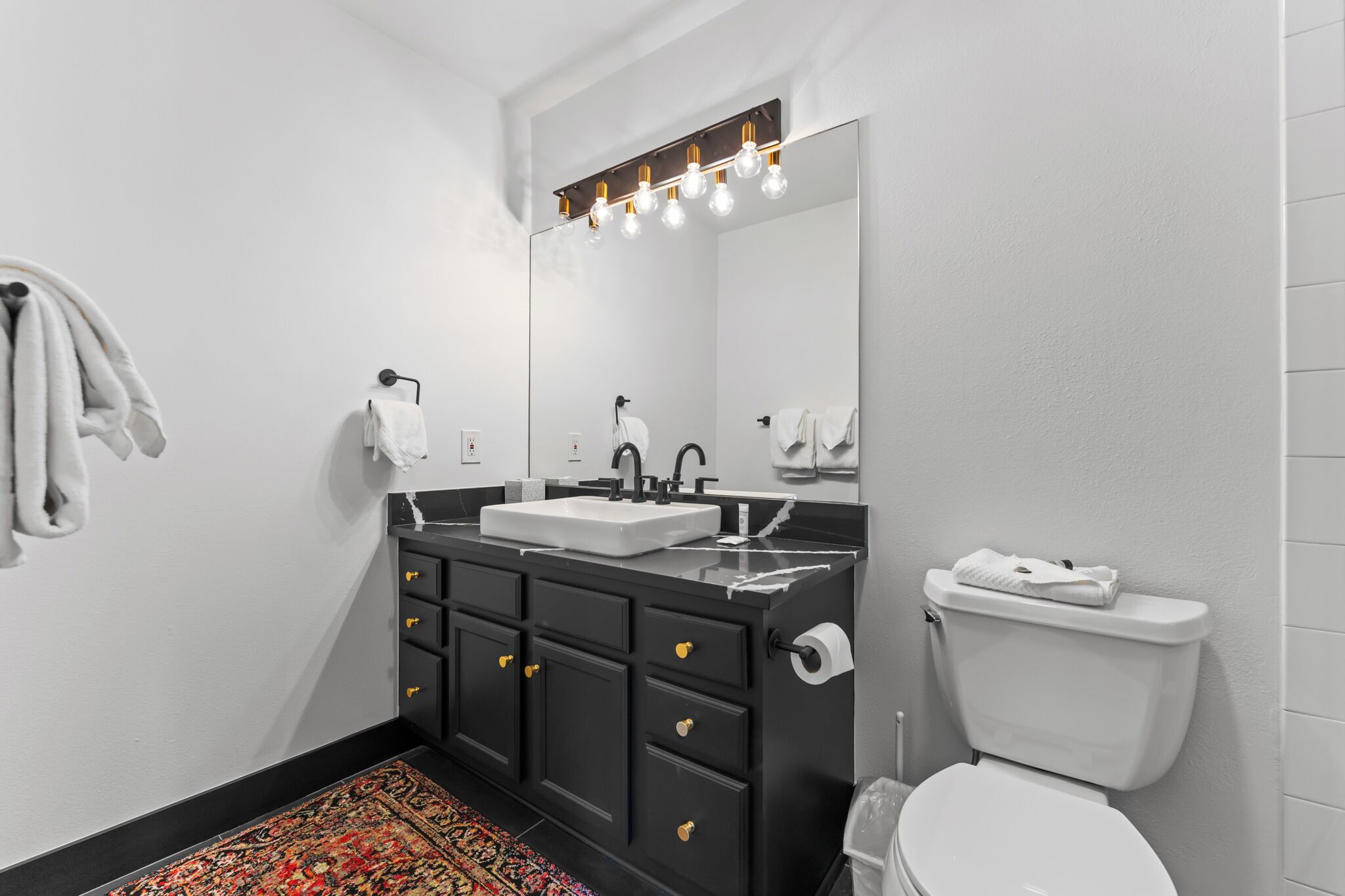Une salle de bain moderne avec un meuble-lavabo noir, un lavabo blanc et des toilettes. Un miroir avec des lumières est placé au-dessus du lavabo, et un tapis à motifs recouvre le sol.