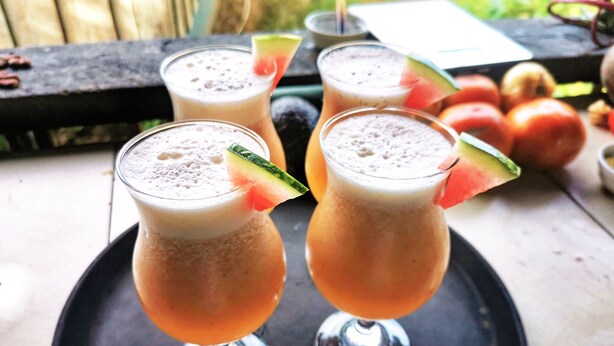 Two glasses of a frothy beverage on a black tray, with a watermelon slice garnish, set on an outdoor table with a wooden bench and a white napkin in the background.