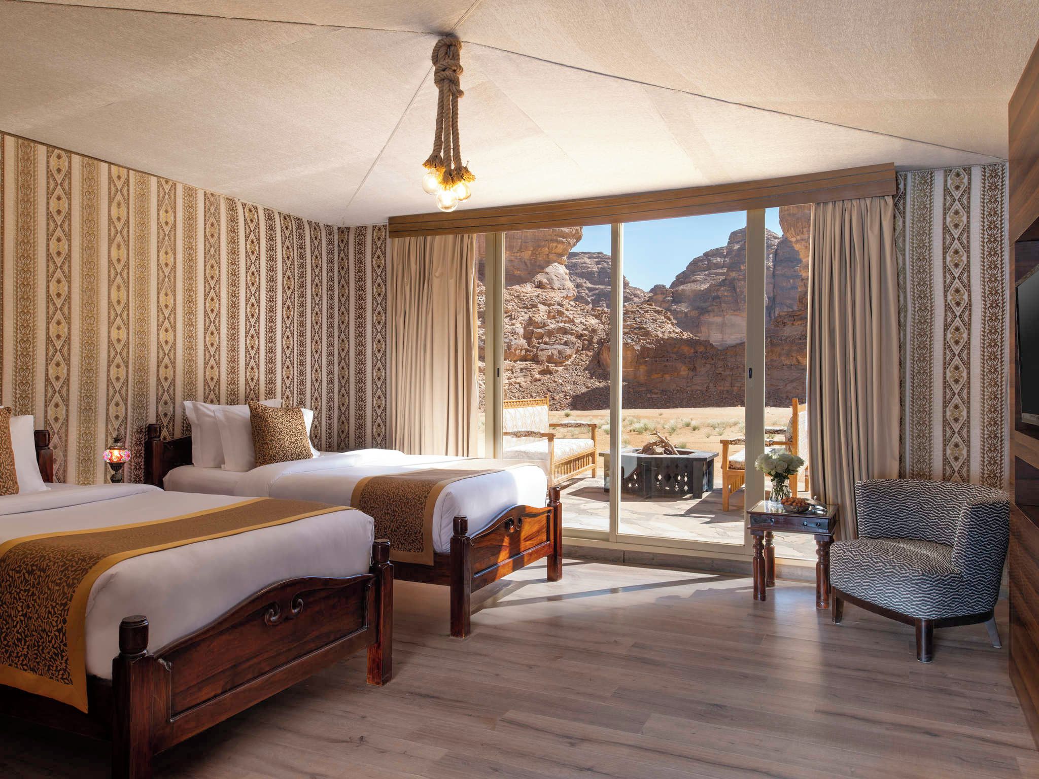 A hotel room with two beds, a large window with a desert view, a wooden floor, and a chandelier.