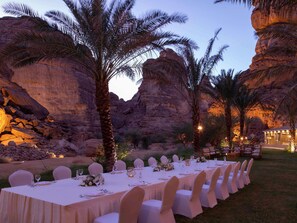 Un área de comedor al aire libre con mesas preparadas para una comida, rodeada de palmeras y formaciones rocosas.