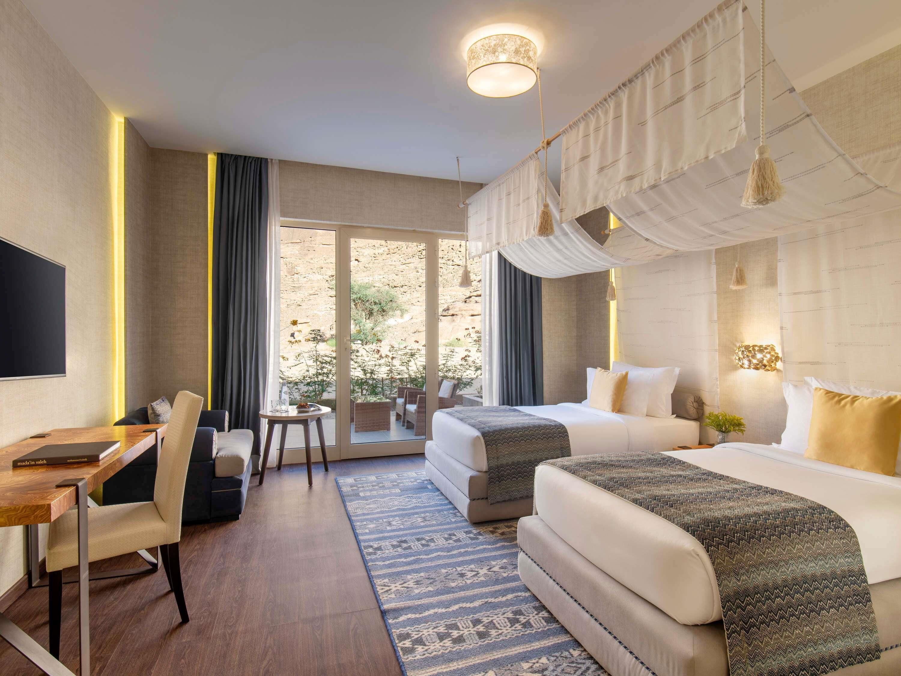 A hotel room with a desk, two beds, a TV, and a large window with curtains.