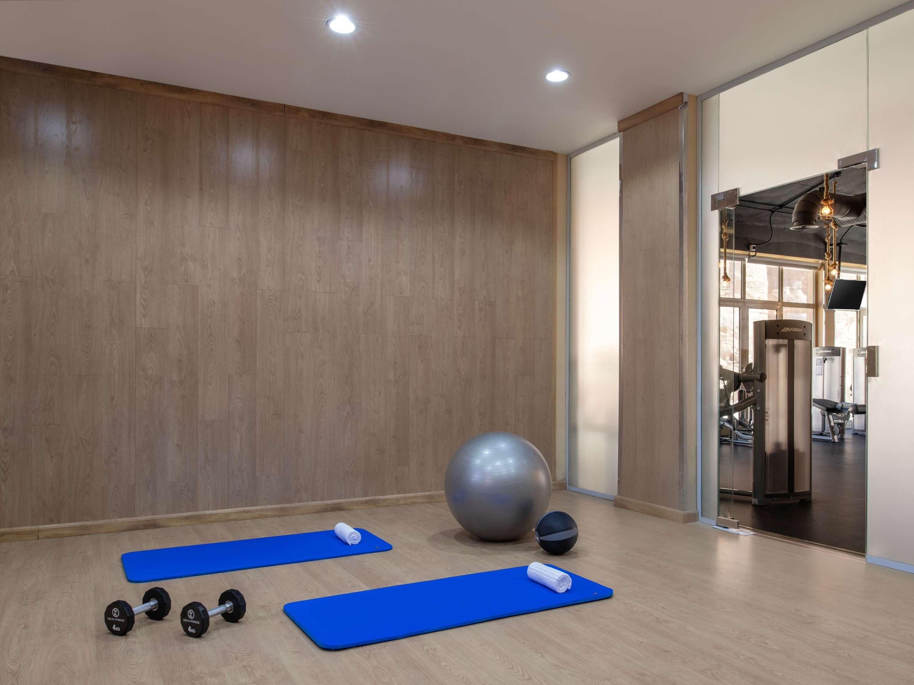Uma academia com colchonetes de exercícios, halteres, uma bola de ginástica grande e uma TV fixada na parede.