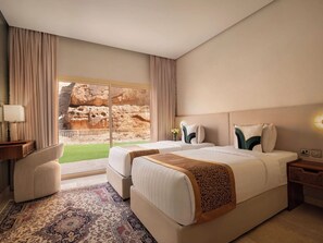Una camera d'albergo con due letti, un angolo salotto, un'ampia finestra vista su formazioni rocciose e un tappeto decorato.