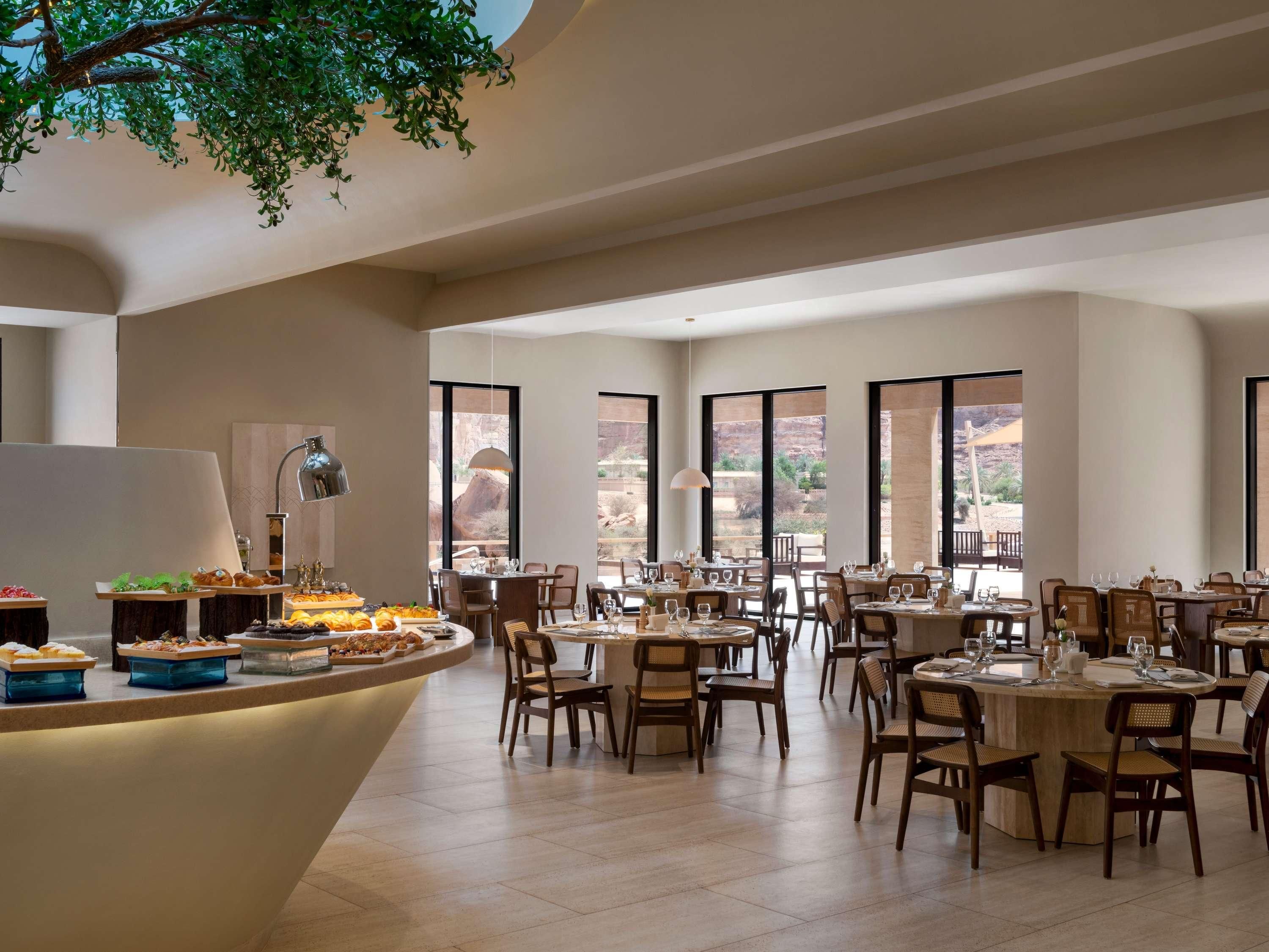 A spacious dining area with multiple tables and chairs, large windows offering a view of the outdoors, and a central buffet counter with various food items.