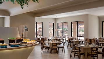 A spacious dining area with multiple tables and chairs, large windows offering a view of the outdoors, and a central buffet counter with various food items.