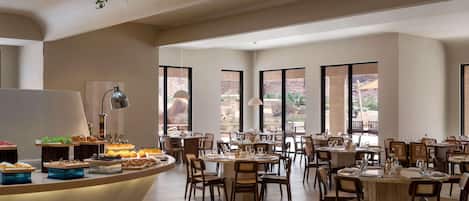 A spacious dining area with multiple tables and chairs, large windows offering a view of the outdoors, and a central buffet counter with various food items.