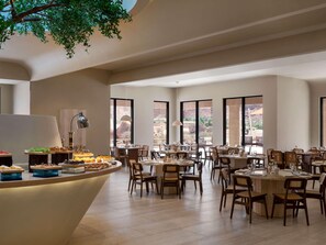 Un comedor amplio con varias mesas y sillas, ventanas grandes que ofrecen vistas al exterior y un mostrador de buffet central con diversos alimentos.