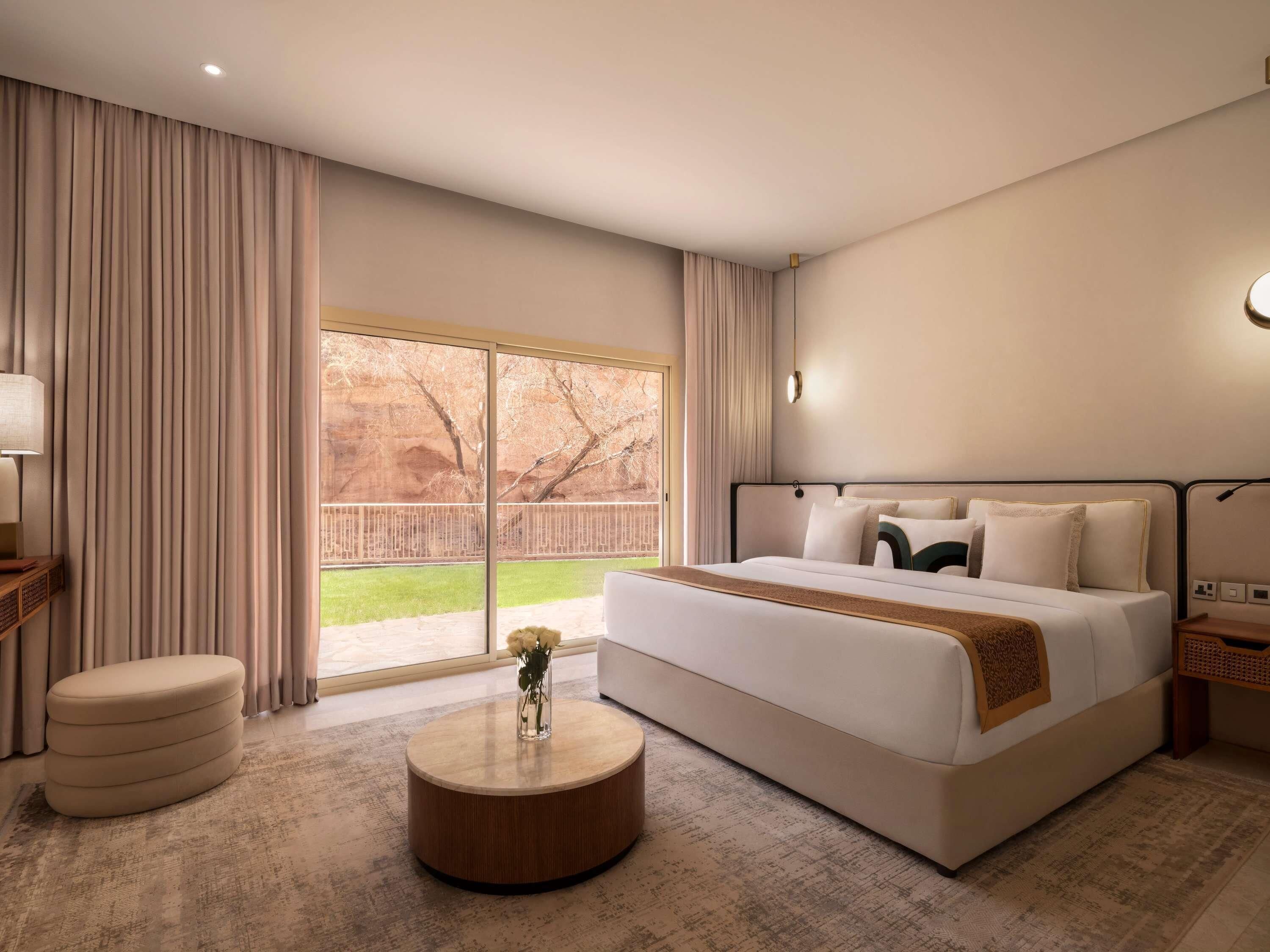 Une chambre d’hôtel avec un grand lit, une table basse ronde, une table de chevet et une vue sur un espace extérieur clos.