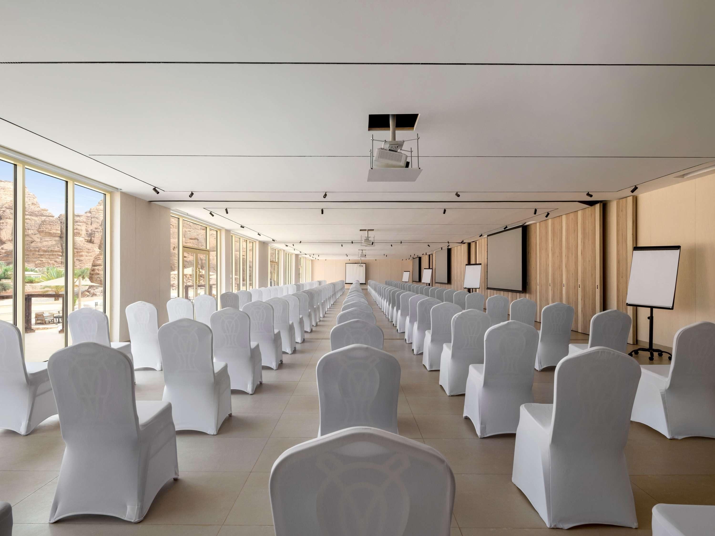 Un salón de conferencias con filas de sillas blancas, ventanas amplias y un proyector en el techo.